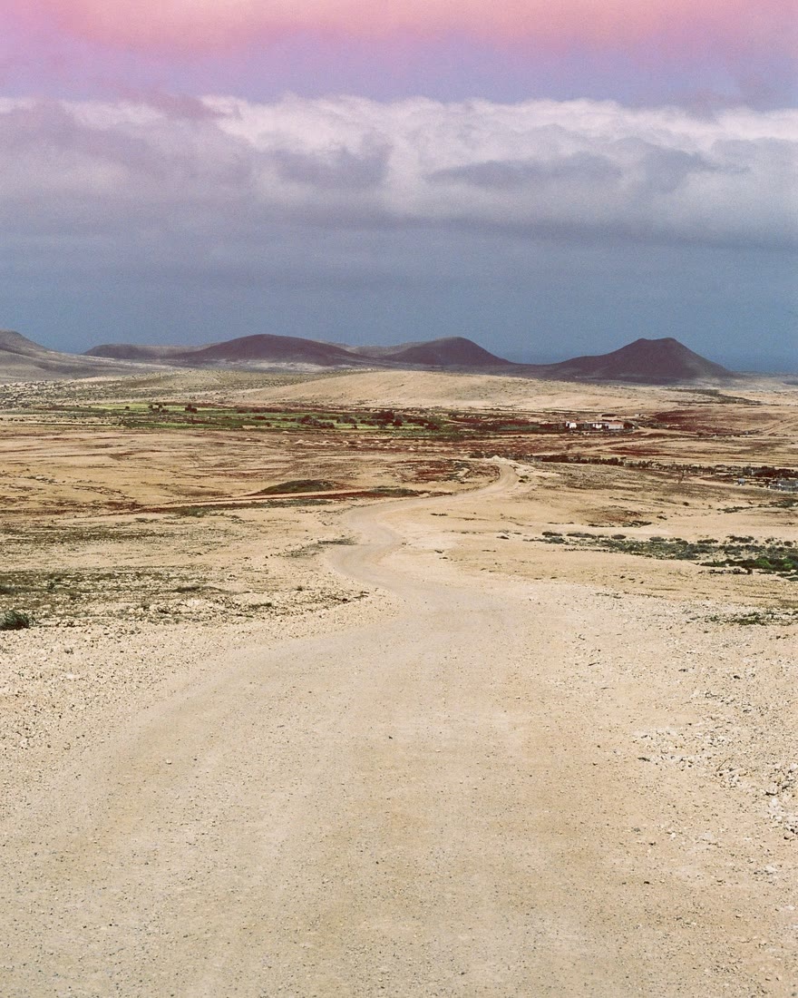 A dirt road winding across a desert toward low volcanic hills, with a soft pink edge fog along the top of the film